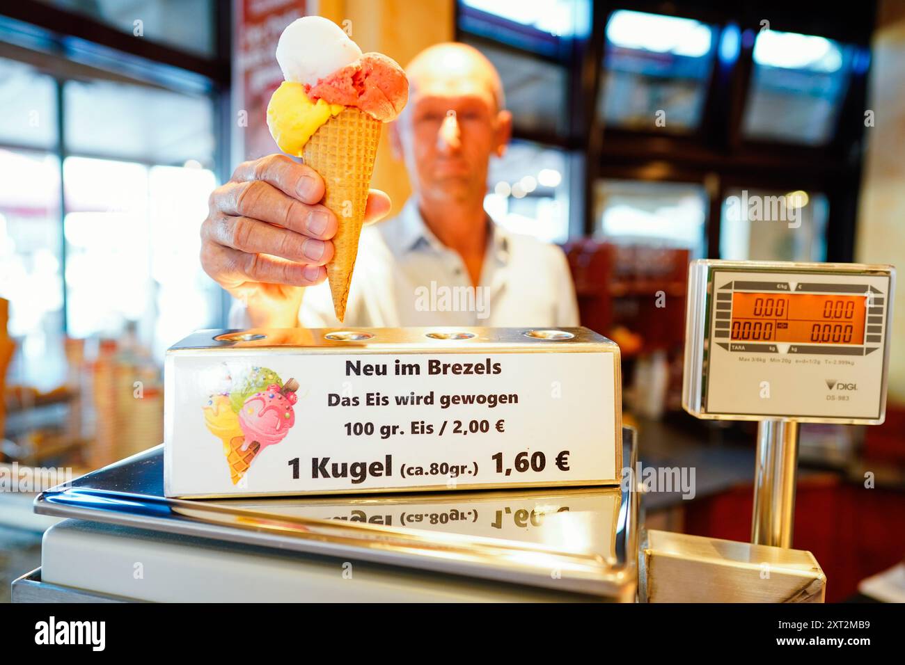 PRODUZIONE - 13 agosto 2024, Baden-Württemberg, Gaggenau: L'operatore Michael Böhmer mette un gelato in un cono sulla bilancia al Café Brezels. Al Café Brezels, nel centro di Gaggenau, il gelato non viene più caricato dalla paletta ma dal grammo. I clienti pagano due euro per 100 grammi di gelato e il prezzo viene adeguato di conseguenza per quantità minori. Foto: Uwe Anspach/dpa Foto Stock