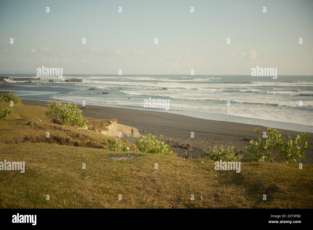 Vista sul mare tropicale con onde del vento dalla cima di una collina erbosa ai margini di una spiaggia di sabbia nera Foto Stock