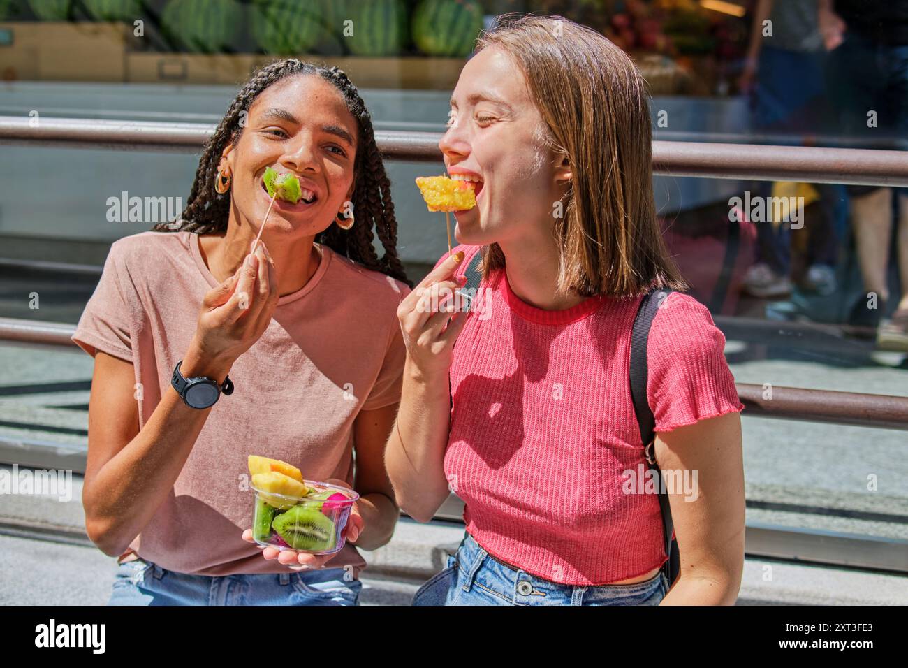 Due amiche multirazziali condividono un momento di gioia mangiando tazze di frutta fresca all'aperto. Stanno sorridendo indicando una forte amicizia e un informale, f Foto Stock