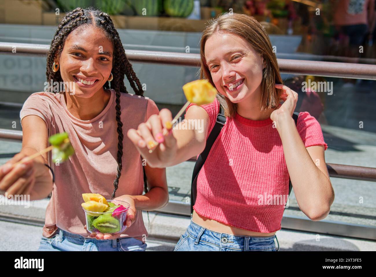 Due donne, una con i capelli intrecciati e l'altra con la parte superiore rossa, condividono con gioia una ciotola di frutta fresca colorata in una giornata di sole che offre la possibilità di guardare la macchina fotografica Foto Stock