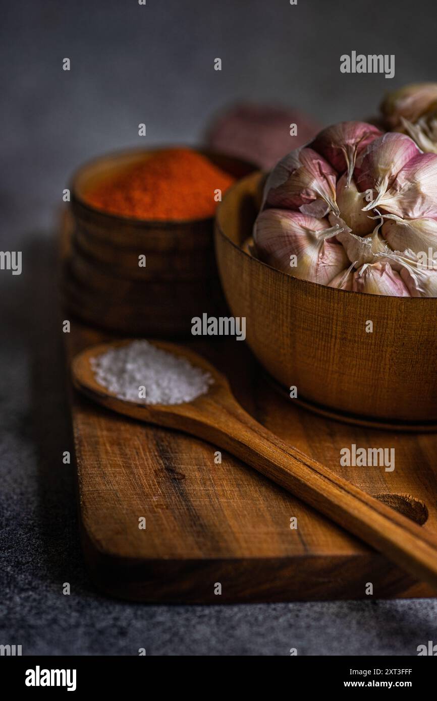 Un'immagine ravvicinata a bassa illuminazione che mostra un bulbo di aglio, spezie e sale disposto ordinatamente su un asse di legno, creando un tema culinario suggestivo Foto Stock