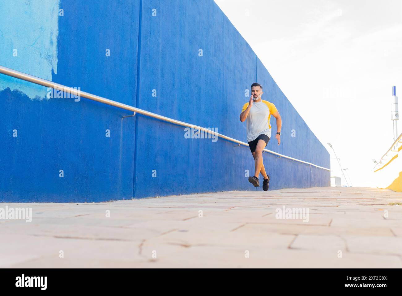 Un uomo indiano in abbigliamento sportivo è visto fare jogging in una giornata di sole, guardando lontano dalla macchina fotografica. Indossa una t-shirt gialla e pantaloncini correndo al fianco Foto Stock