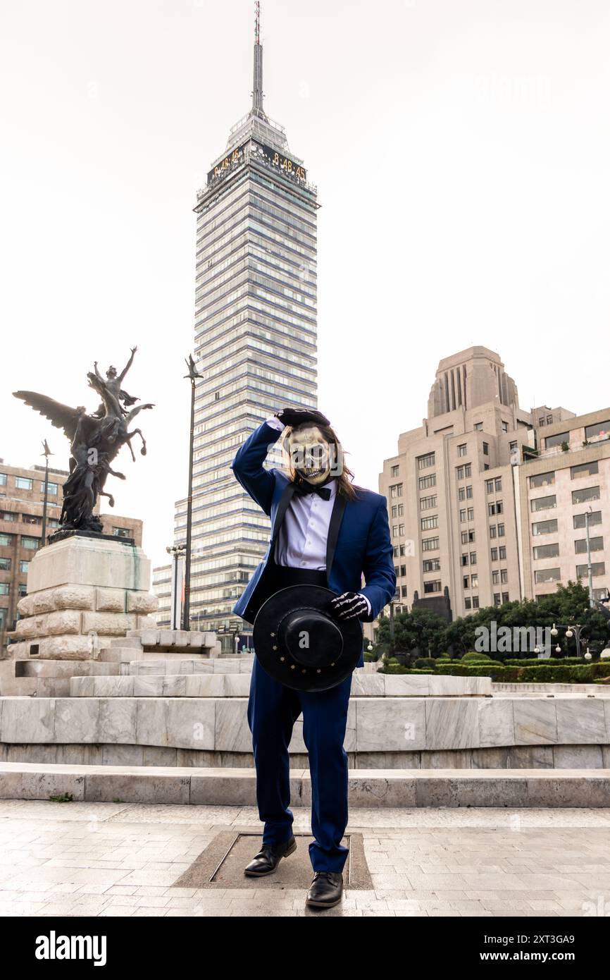 Un uomo con una tuta formale e una maschera cranica di Catrin si posa di fronte all'iconica Torre Latinoamericana a città del Messico, simboleggiando la festa del giorno dei morti Foto Stock