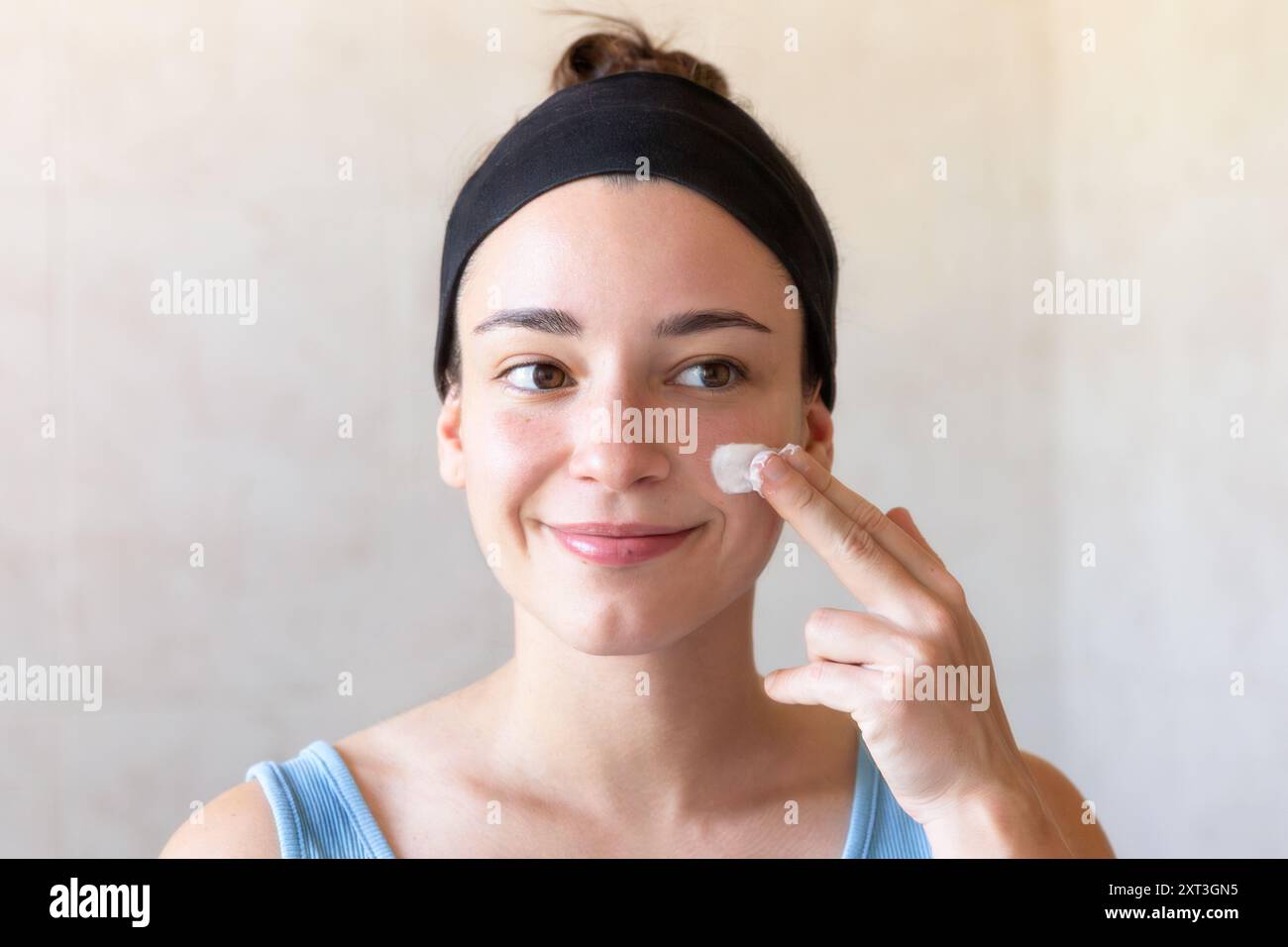 Una giovane donna con una canotta blu, che le applica una crema idratante sul viso, si trova di fronte a uno sfondo neutro, incarnando una routine quotidiana di cura di sé Foto Stock