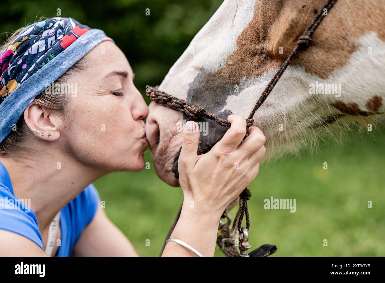 Vista ravvicinata di una donna che baciava affettuosamente un cavallo dipinto sulla museruola, in piedi in un pascolo verde vivido. Il momento attivo e tenero captu Foto Stock