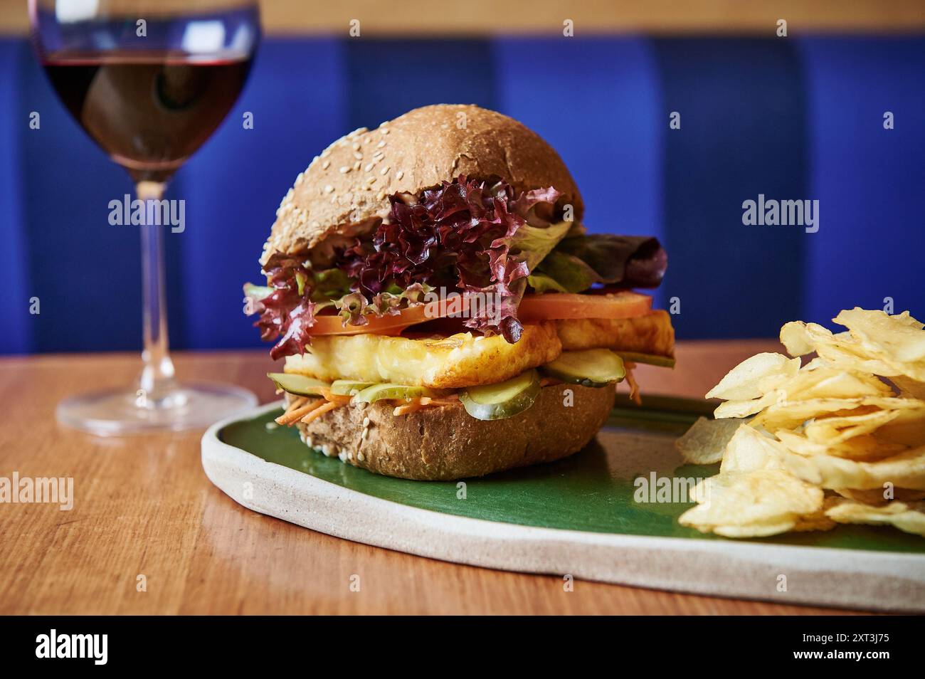 Un delizioso halloumi hamburger servito con patatine croccanti e un bicchiere di vino rosso, presentato su un piatto di ceramica moderna sullo sfondo blu. Foto Stock