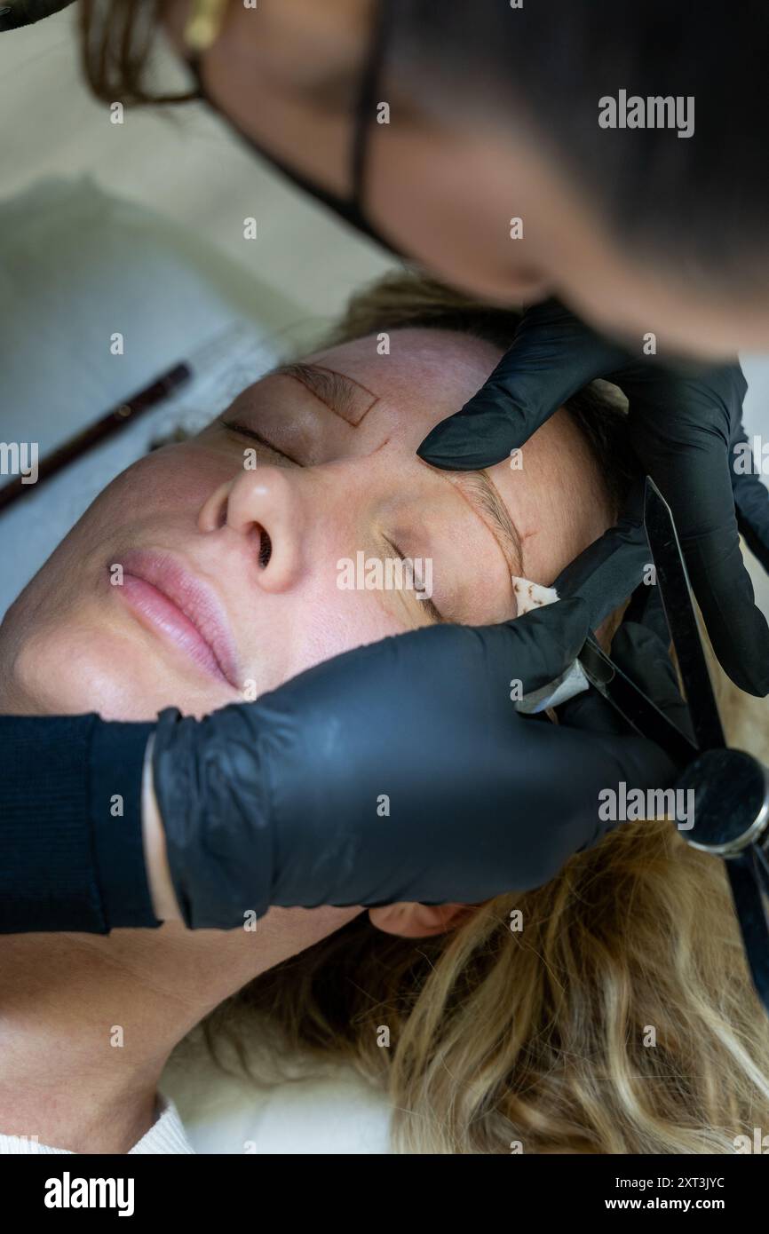 Un estetista esperto che indossa guanti neri disegna e misura attentamente le sopracciglia del cliente per garantire una microblad precisa e geometricamente bilanciata Foto Stock