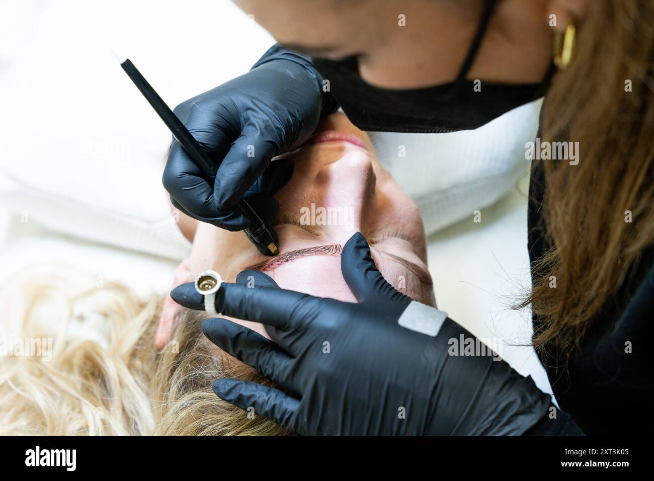Un estetista esperto esegue con attenzione una procedura di microblading sulle sopracciglia di un cliente, migliorandone lo spessore e la forma in un ambiente clinico. Foto Stock