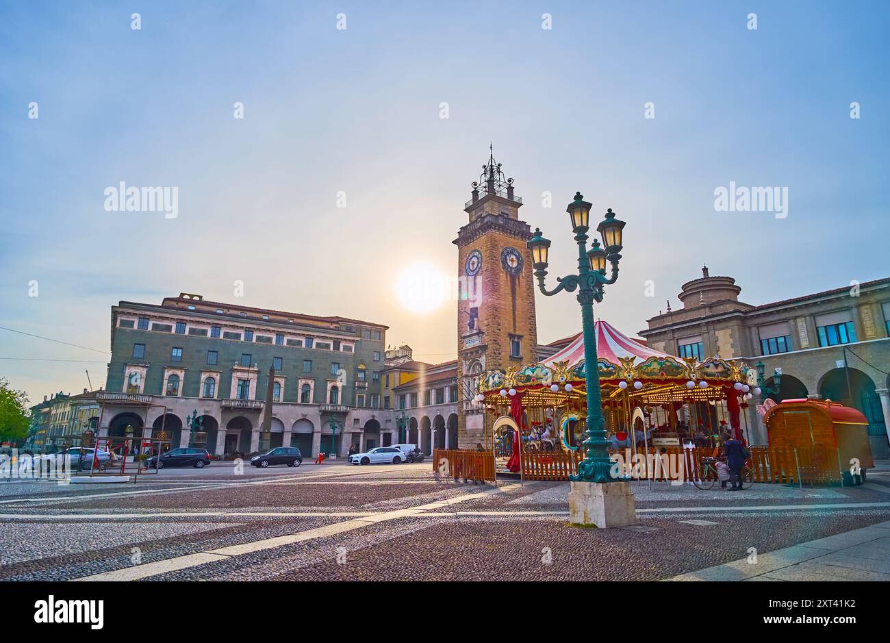 Il tramonto nebbioso su Piazza Vittorio Veneto con lampioni vintage, giostra e Torre commemorativa sullo sfondo, Bergamo, Italia Foto Stock