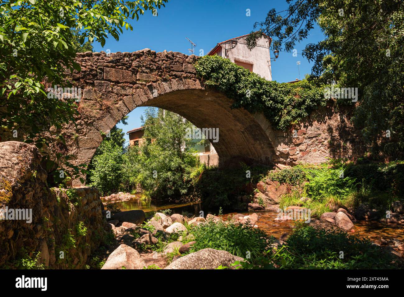 Uno storico ponte in pietra si arrocca graziosamente su un tranquillo ruscello a Hervas, Caceres. Circondato da vegetazione lussureggiante e bagnato dalla luce del sole, si trova a circa Foto Stock