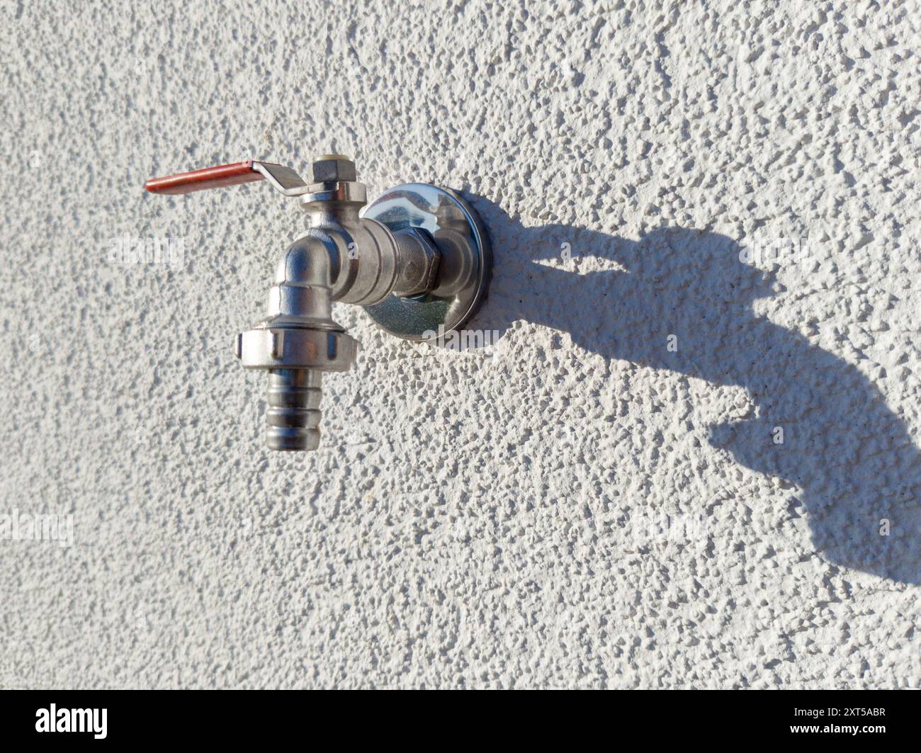 Primo piano di un rubinetto in acciaio inossidabile fissato a una parete ruvida e ruvida che crea un'ombra forte. Foto Stock