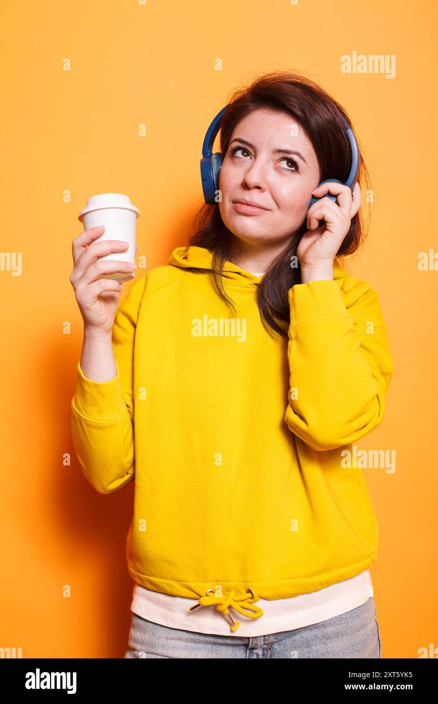 Calma donna caucasica che indossa le cuffie mentre si diverte a divertirsi con la sua tazza di caffè. Una bruna simpatica e rilassata che ascolta la musica sulla piattaforma di streaming audio utilizzando le cuffie wireless. Foto Stock