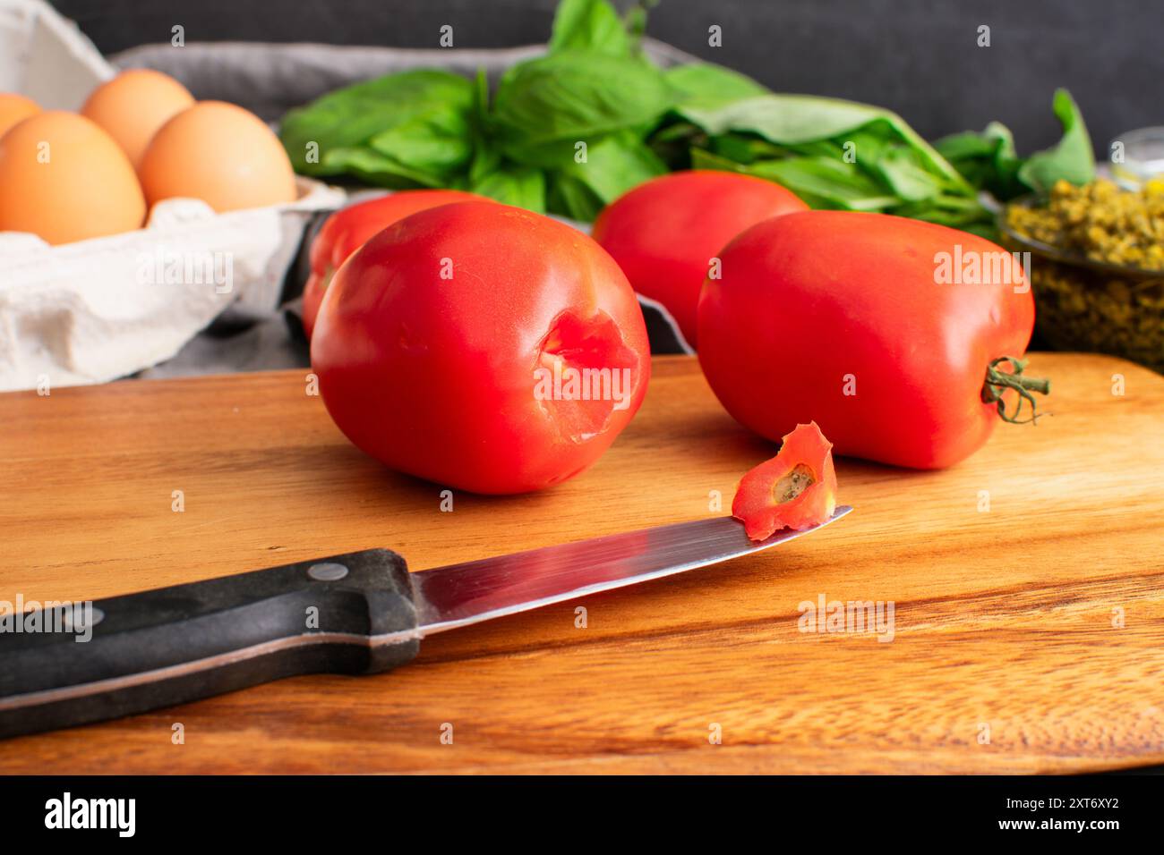 Pomodoro Roma carotato su un tagliere di legno: Pomodori prugna e coltello da cucina su un tavolo di legno con uova, basilico e un piatto di pesto sullo sfondo Foto Stock