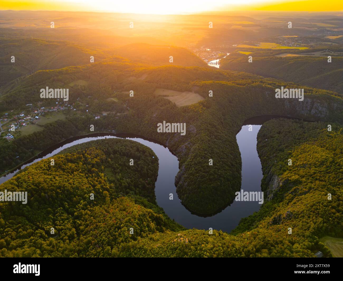Il fiume Moldava scorre graziosamente attraverso lussureggianti colline verdi mentre il sole tramonta, gettando un bagliore dorato sul paesaggio della Cechia. Questa tranquilla scena cattura l'essenza dell'estate. Foto Stock