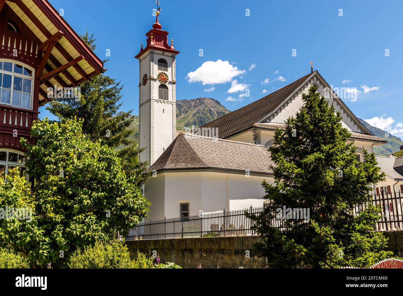 La chiesa parrocchiale di San Pietro e Paolo, con il suo campanile rosso e la torre dell’orologio, domina il paesaggio urbano di Andermatt, un fianco montuoso boscoso che si innalza dietro di essa. La chiesa fu costruita nel 1601/1602 sotto la guida del mastro costruttore Johannes Schmid di Hospental, ampliata nel 1694 e ristrutturata in stile neobarocco nel 1903–1905; è considerata una delle chiese più importanti del cantone di Uri. Bahnhofstrasse, Andermatt, Uri, Svizzera. Foto Stock