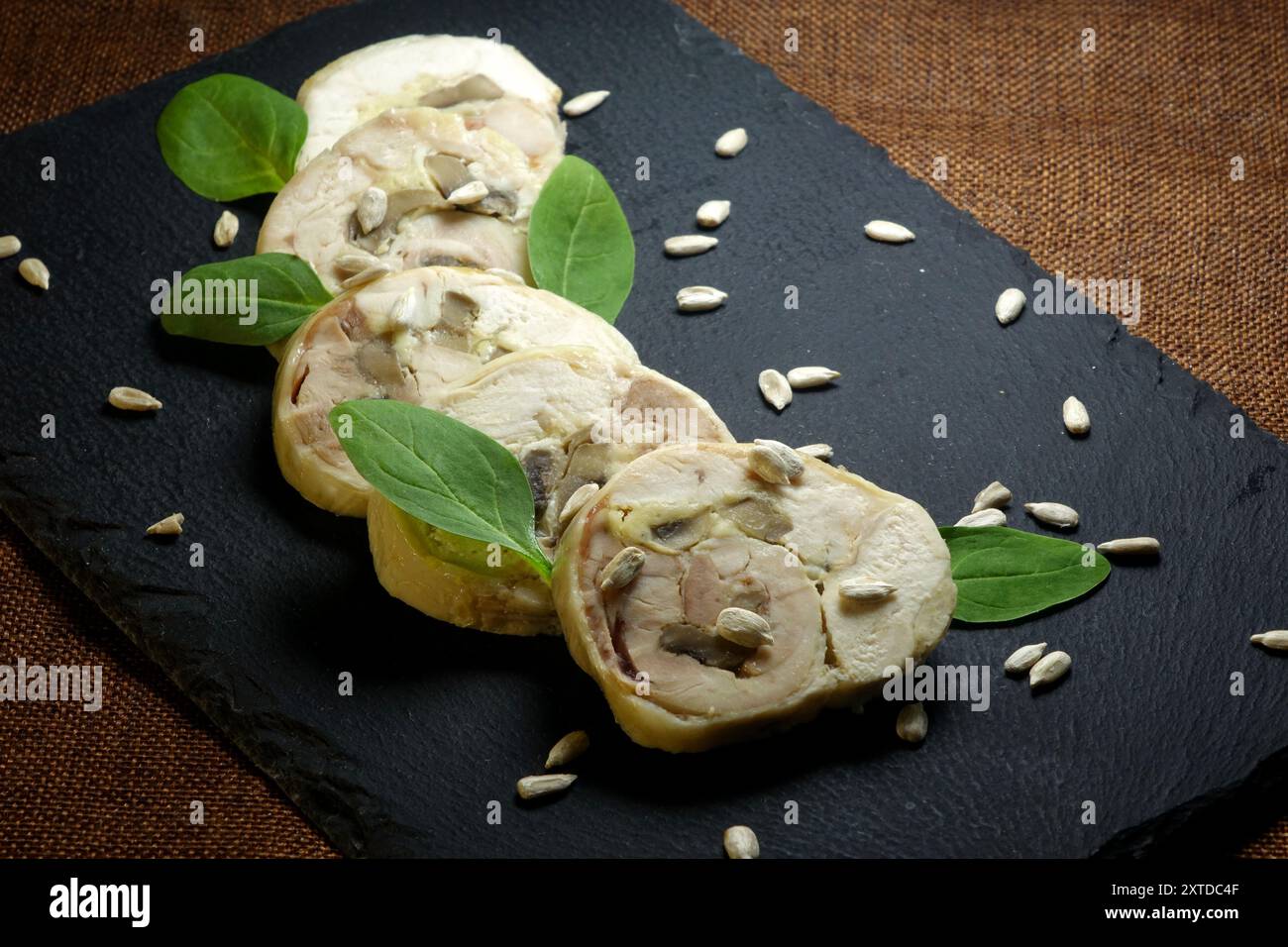 Le fette di un involtino di carne sono elegantemente disposte su un bordo di ceramica nera, accentuate da foglie di spinaci freschi e semi di girasole sparsi. Foto Stock