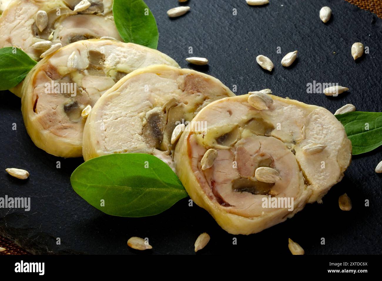 Il rotolo di carne a fette è disposto artisticamente su un pannello di ceramica nera, arricchito da foglie di spinaci freschi e semi di girasole sparsi. Foto Stock