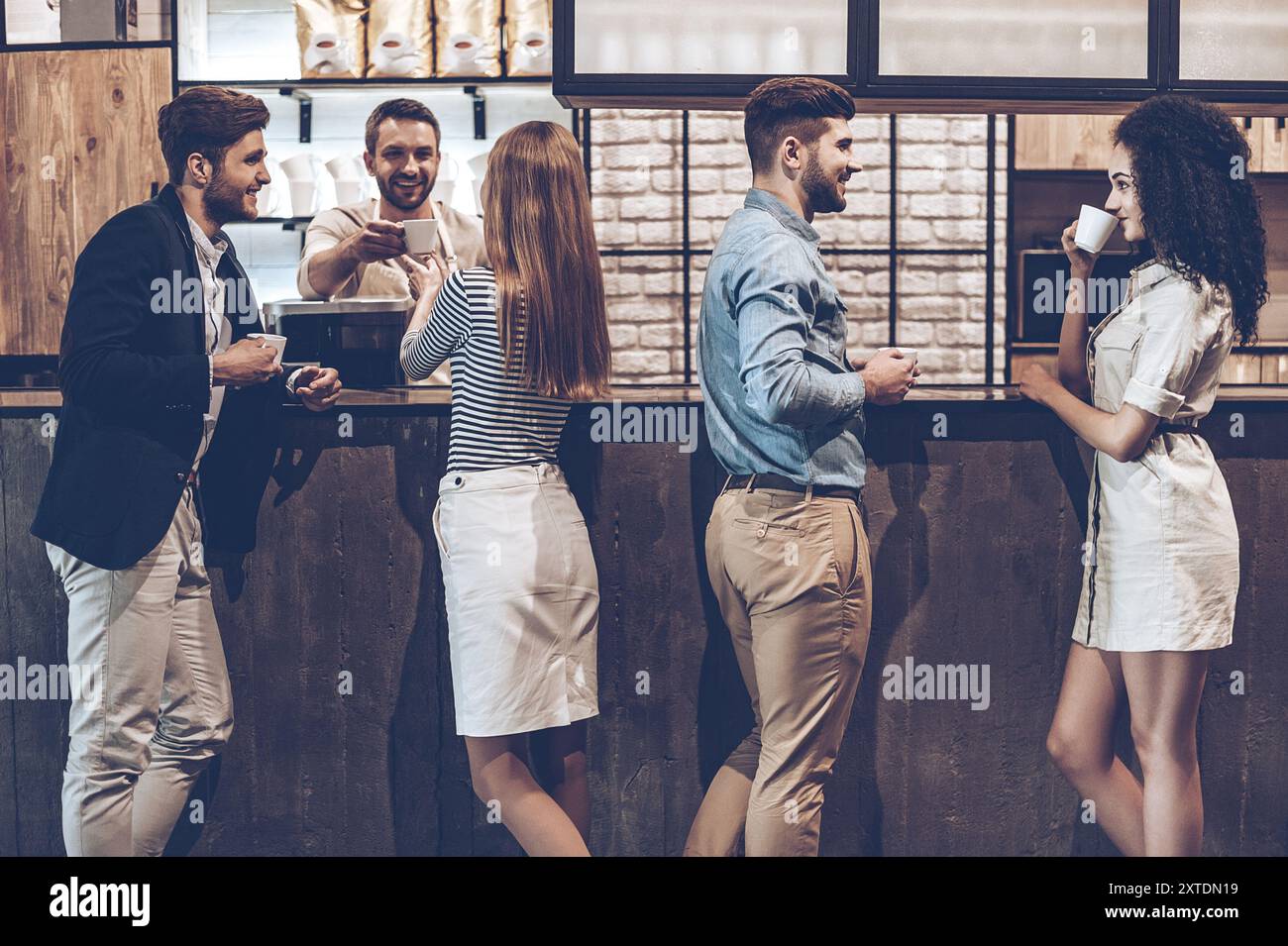Vibrazioni mattutine nel caffè. Gruppo di giovani che bevono caffè e discutono di qualcosa mentre si trovano al bancone del bar Foto Stock