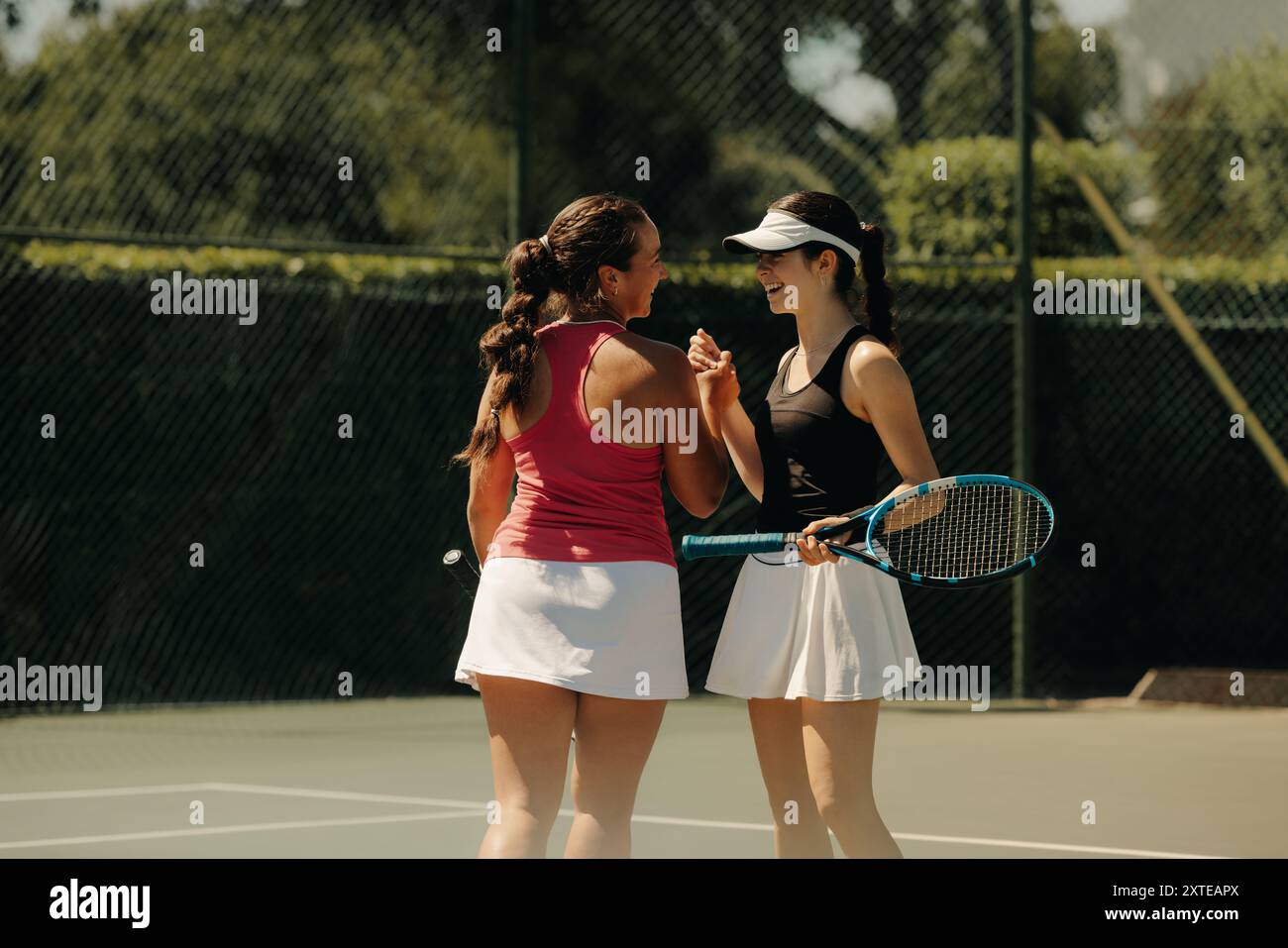 Due donne che conversano e sorridono durante una giornata di sole sul campo da tennis tenendo racchette e mostrando cameratismo. Foto Stock