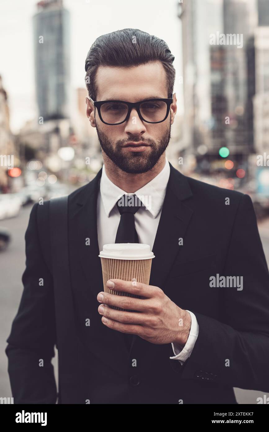 Gusta il caffè in movimento. Giovane sicuro di sé in completo completo possesso di una tazza di caffè e guarda la fotocamera mentre si trova all'aperto con il paesaggio urbano nel bac Foto Stock