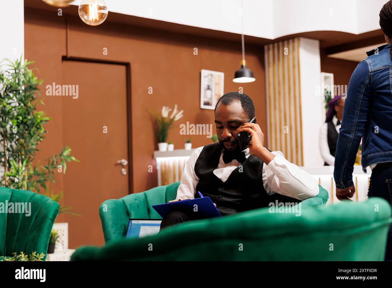Un fattorino afroamericano dell'hotel seduto nella hall a parlare al telefono e a prendere appunti. Il portiere chiama il taxi contattando il servizio di trasporto, assistendo gli ospiti nell'organizzazione del trasporto Foto Stock