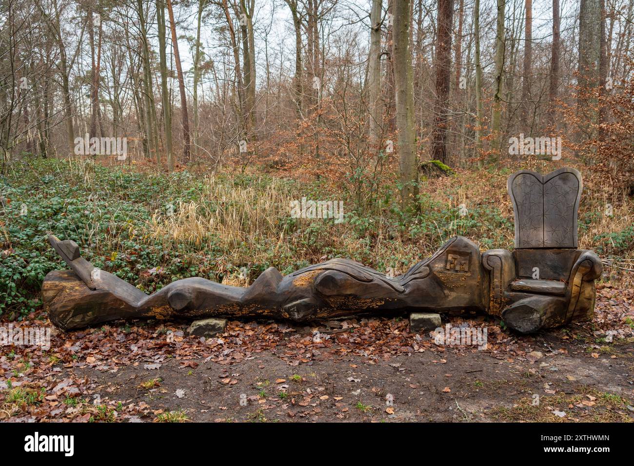 Una lunga panca in tronco intagliato culmina in una poltrona in legno scolpita in modo ornato, creando un'installazione seduta capricciosa nella foresta. Foto Stock