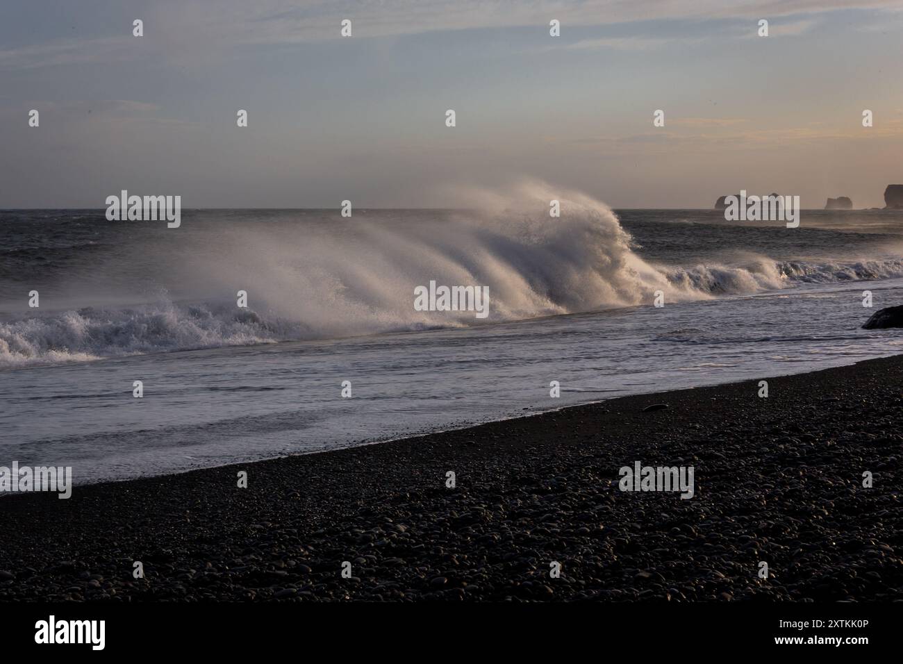 Le onde delle sneaker si infrangono sulla costa di Reynisfjara Black Sand Beach, con forti venti che soffiano acqua e sabbia nera vulcanica, tramonto, Islanda. Foto Stock