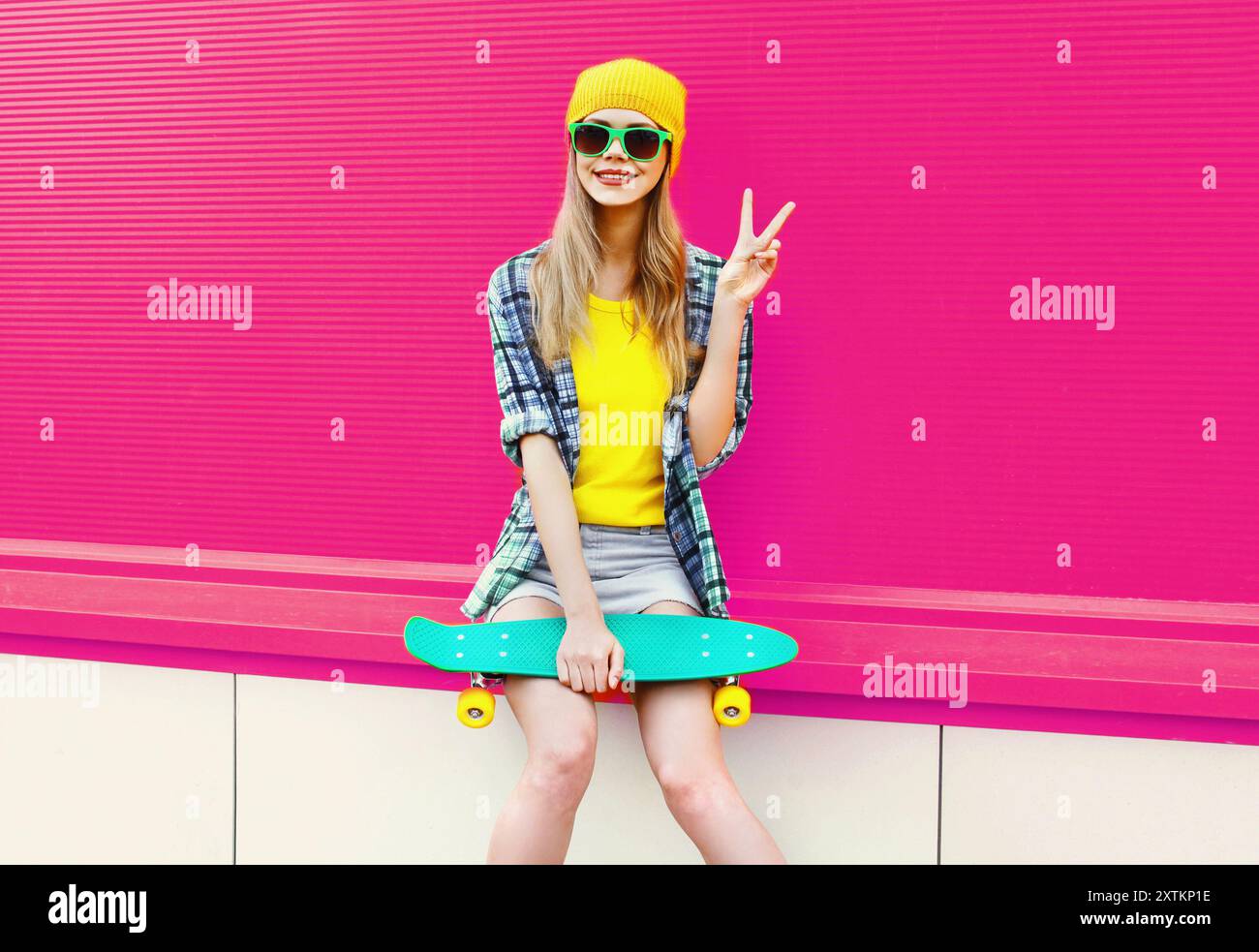 Elegante donna sorridente con skateboard che mostra un gesto di pace con un cappello giallo colorato su sfondo rosa Foto Stock