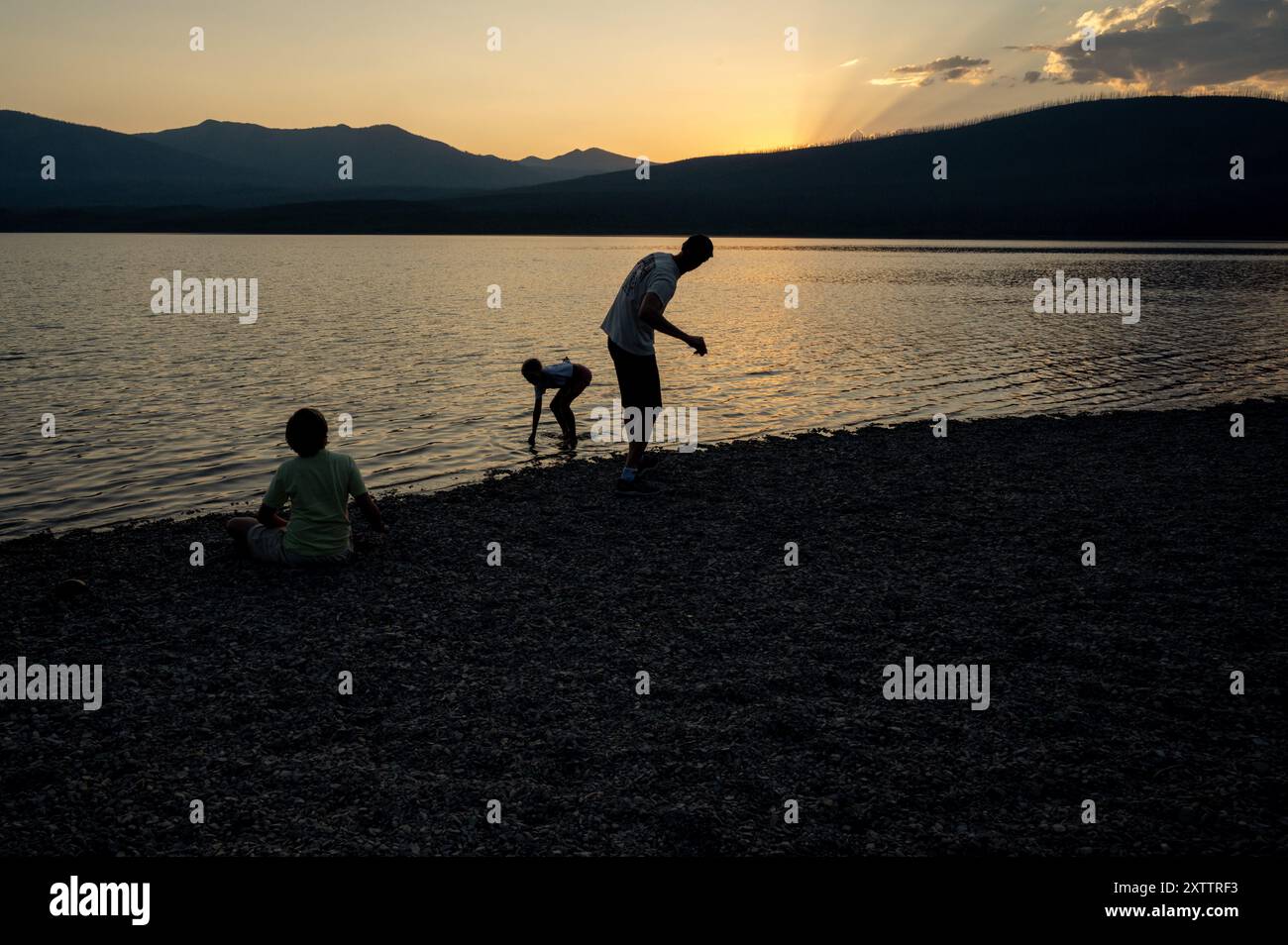 Silhouette di famiglia in piedi in un lago al tramonto saltando rocce Foto Stock