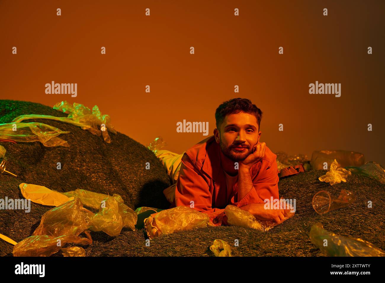 Un uomo giace in un letto di plastica, circondato da oggetti scartati, fissando il cielo arancione. Foto Stock