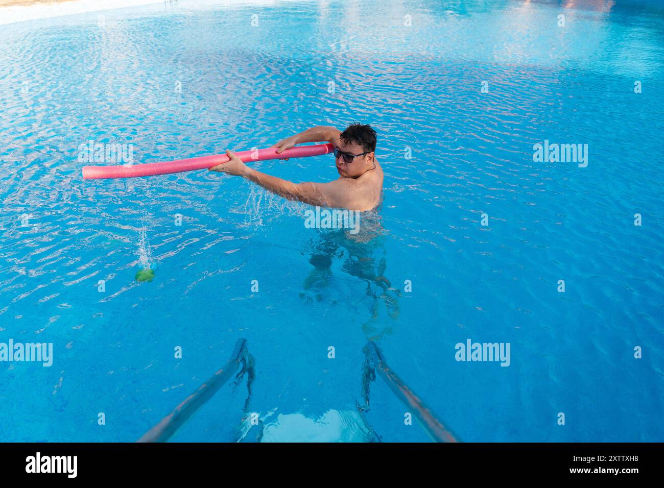 Un uomo con gli occhiali da sole muove giocosamente un noodle rosa in una piscina blu, creando uno spruzzo. Gli piace un momento libero e attivo in acqua Foto Stock