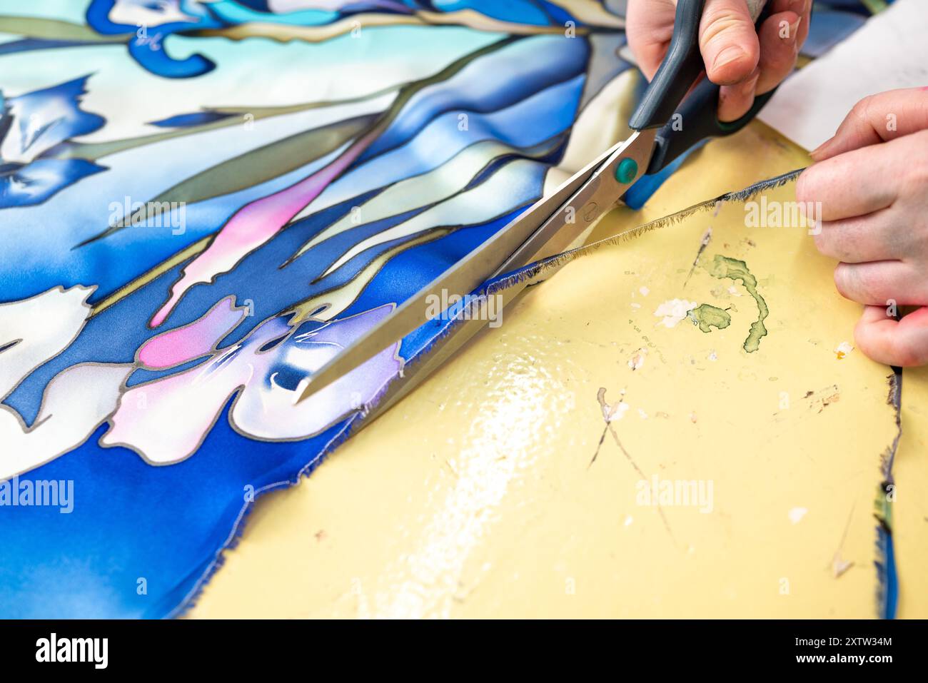 Mani che tagliano con cura intricato tessuto floreale blu e verde con forbici argentate su un tappetino da taglio giallo. La precisione e il dettaglio del taglio p Foto Stock