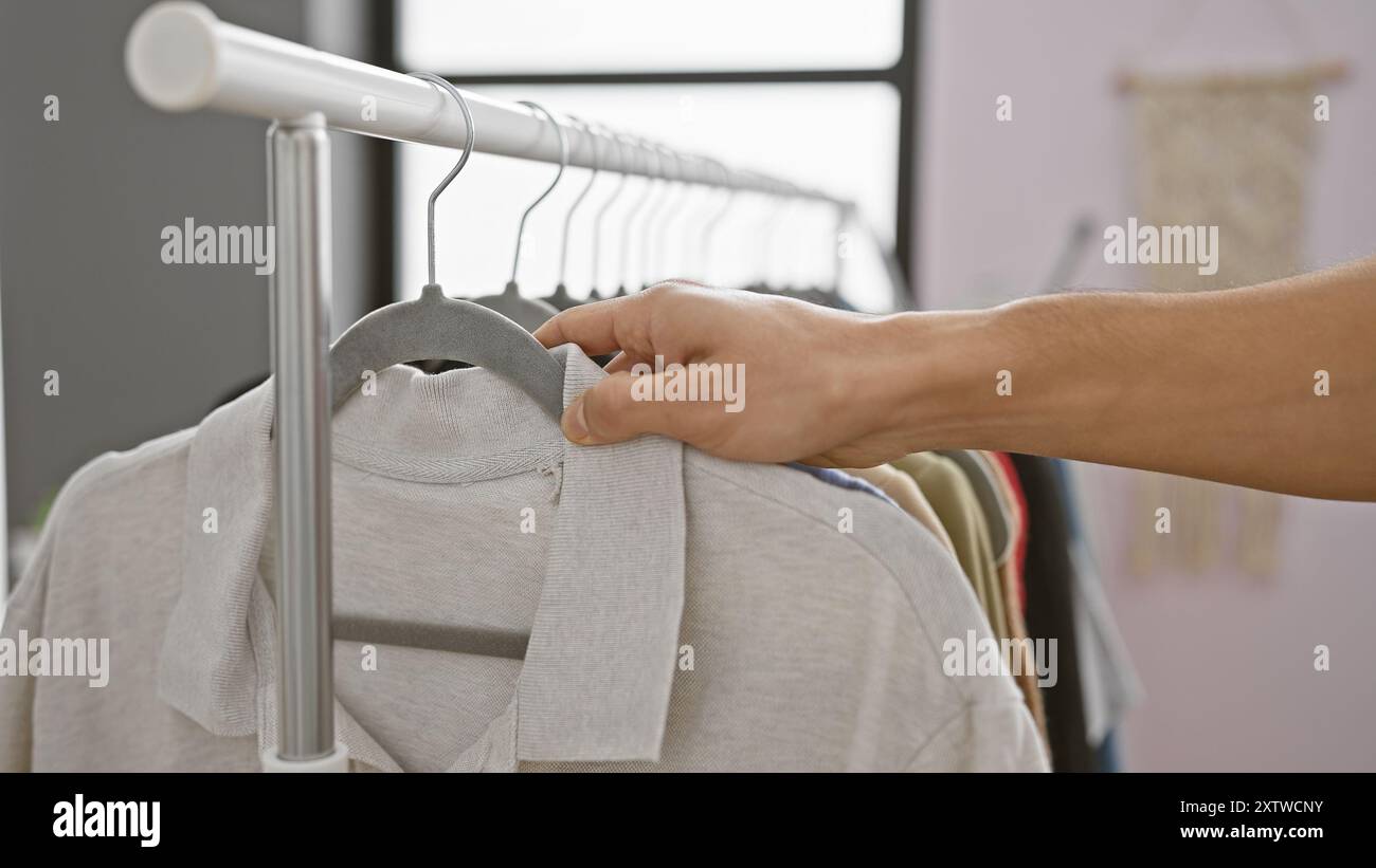 La mano di un uomo seleziona una camicia da un guardaroba in una stanza ben illuminata, simboleggiando le scelte di moda e abbigliamento quotidiano. Foto Stock