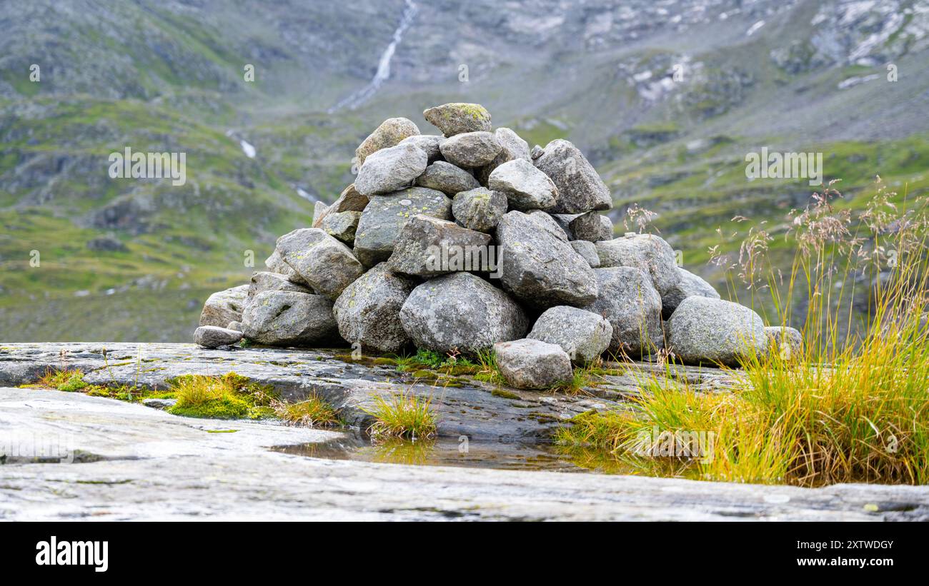 Un cairn accuratamente impilato sorge su un terreno roccioso con vegetazione lussureggiante e sfondo di una cascata. Foto Stock