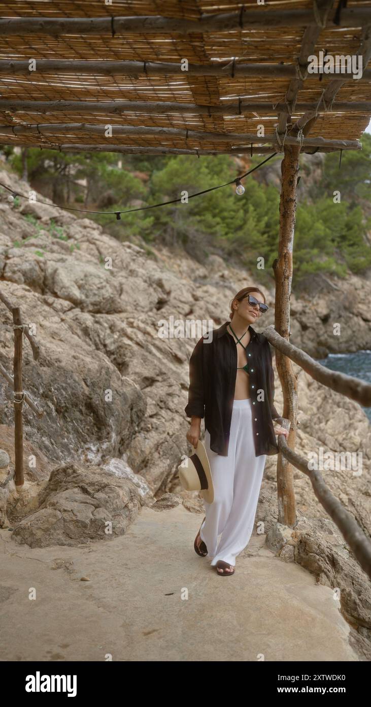 Donna che si rilassa all'aperto a cala deia a maiorca, spagna, indossa occhiali da sole e indossa un cappello, circondata da un terreno roccioso e da vegetazione sotto un rustico Foto Stock