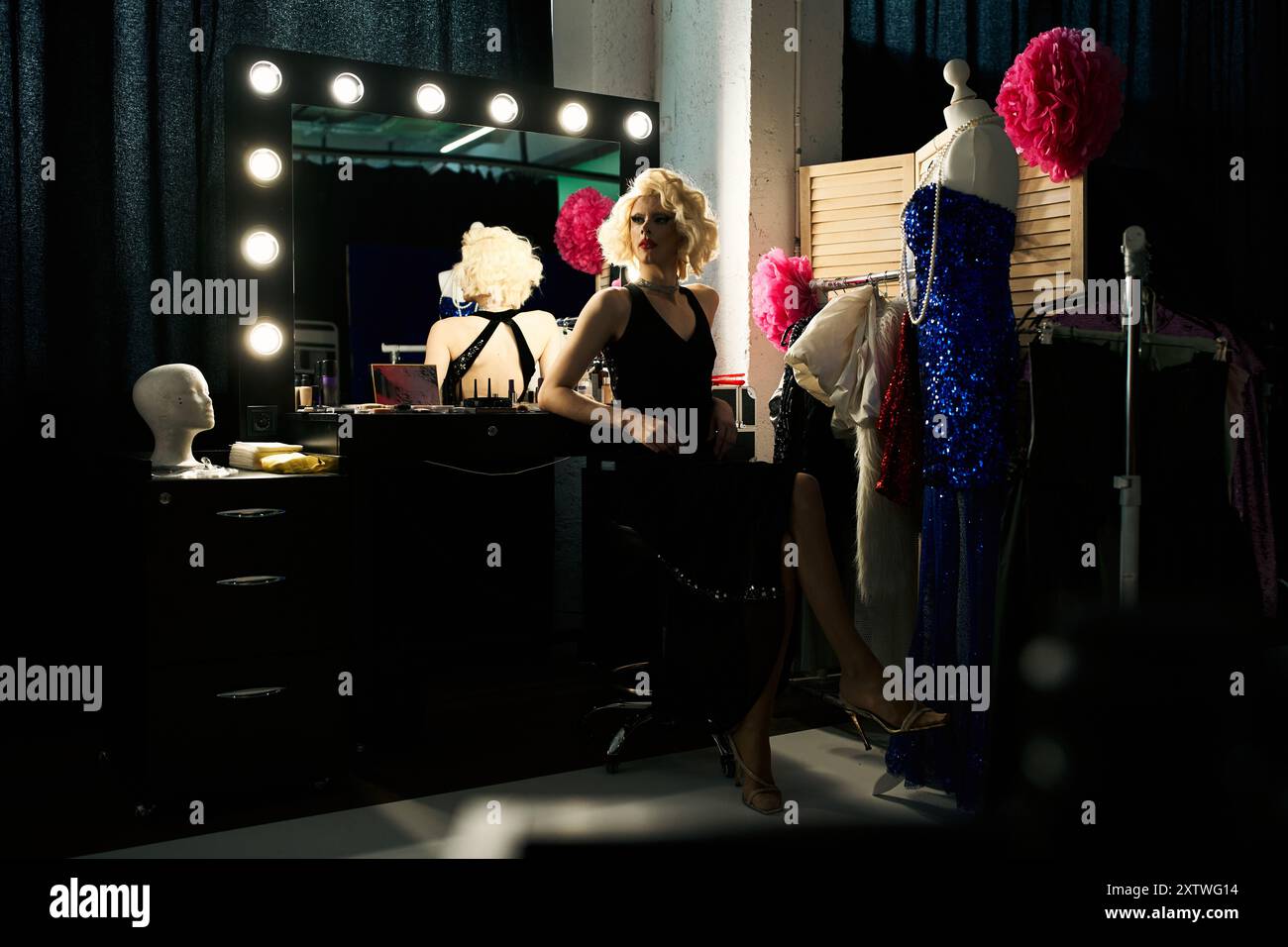 Una drag queen siede ad un tavolo da spogliatoio, preparandosi per una performance, con un costume su un manichino nelle vicinanze. Foto Stock Una drag queen siede ad un tavolo da spogliatoio, preparandosi per una performance, con un costume su un manichino nelle vicinanze. Foto Stock