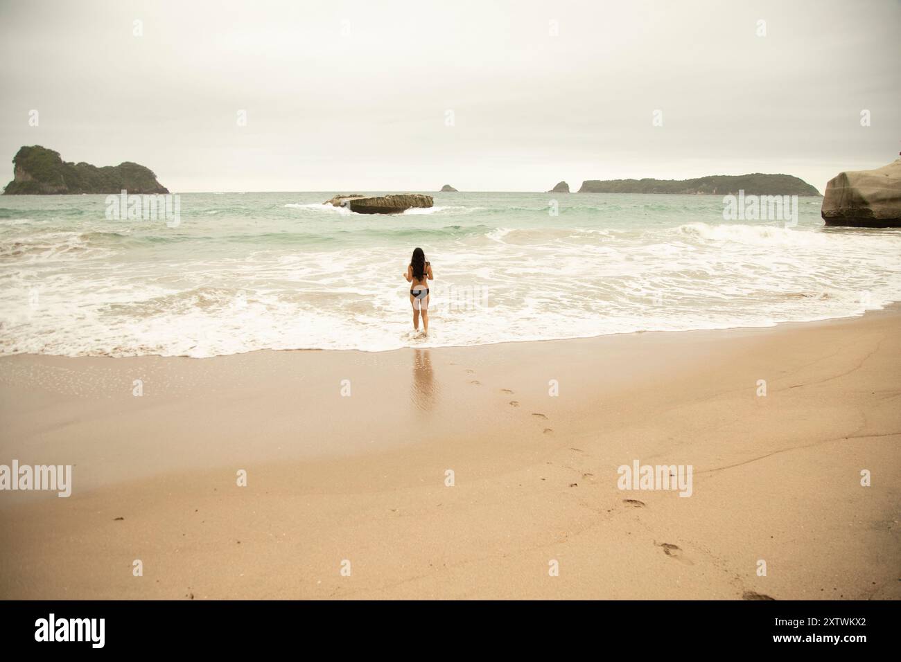 Una donna sta con le spalle alla telecamera, guardando le onde dell'oceano e le formazioni rocciose lungo una spiaggia tranquilla. Foto Stock