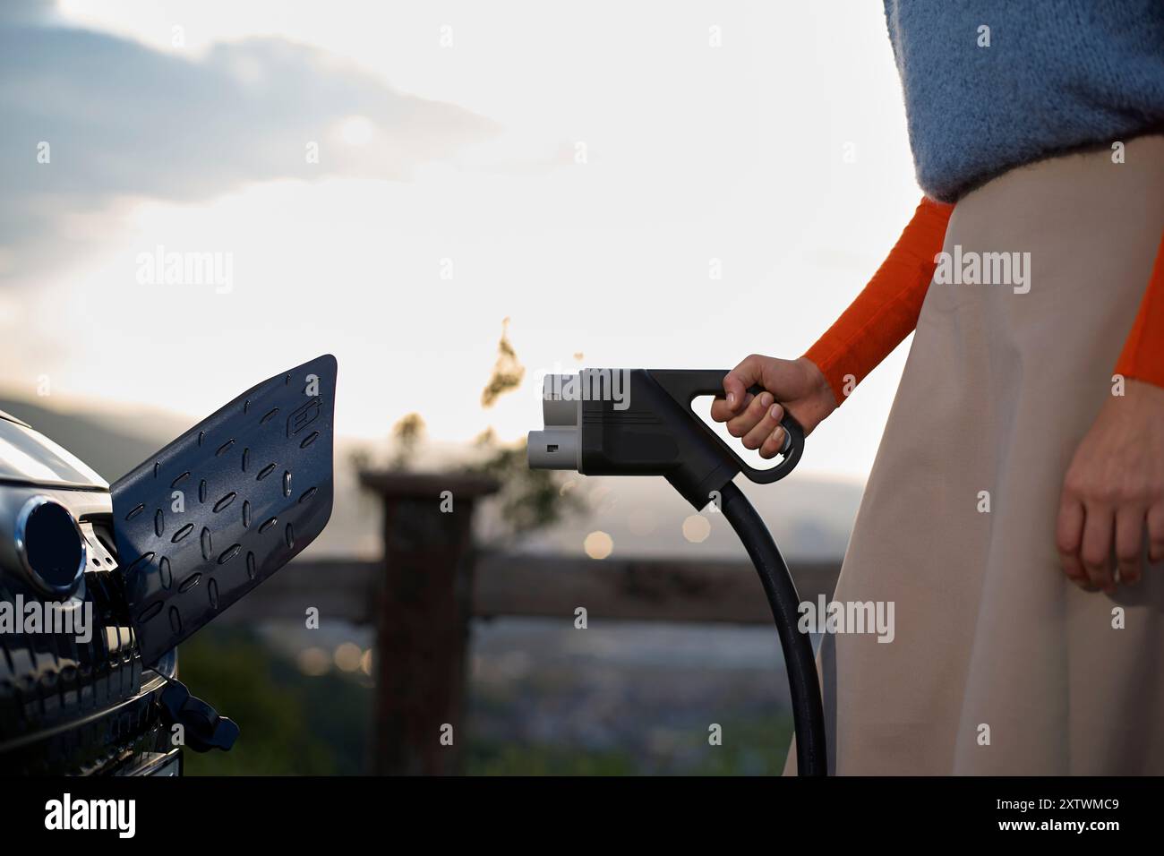 Primo piano della mano di una persona che collega un caricabatterie per auto elettriche a una stazione di ricarica durante il tramonto. Foto Stock