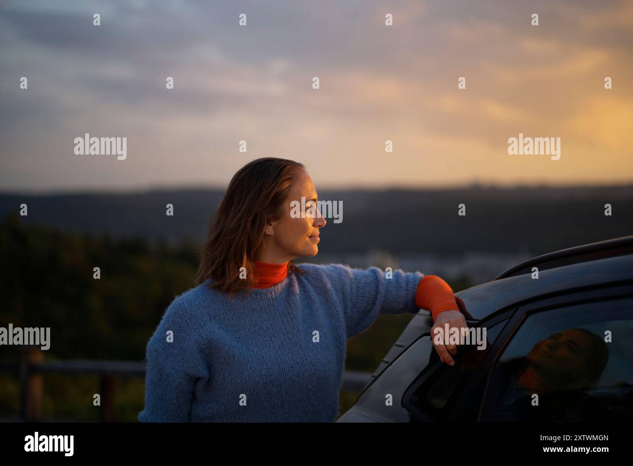 Donna appoggiata su un'auto che gode di una vista panoramica del tramonto. Foto Stock