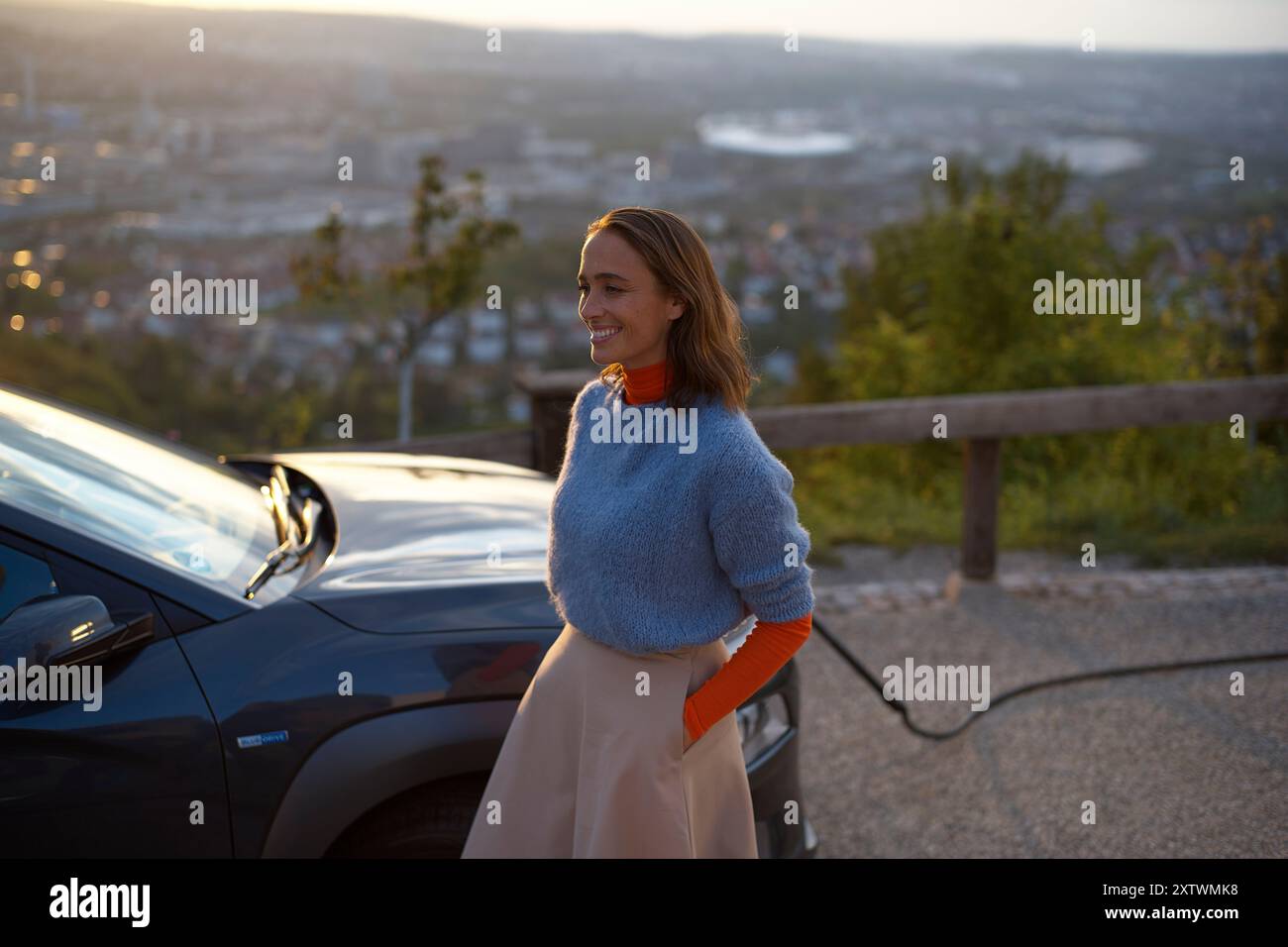 Donna sorridente in piedi accanto a un'auto con un libro arancione, con un paesaggio urbano panoramico sullo sfondo durante il tramonto. Foto Stock