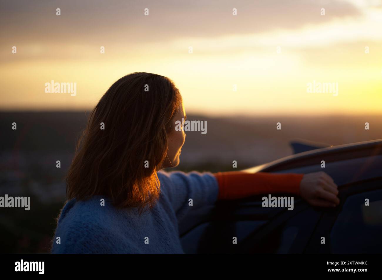 Donna appoggiata sulla sua auto che gode di una vista panoramica del tramonto da un alto punto panoramico. Foto Stock