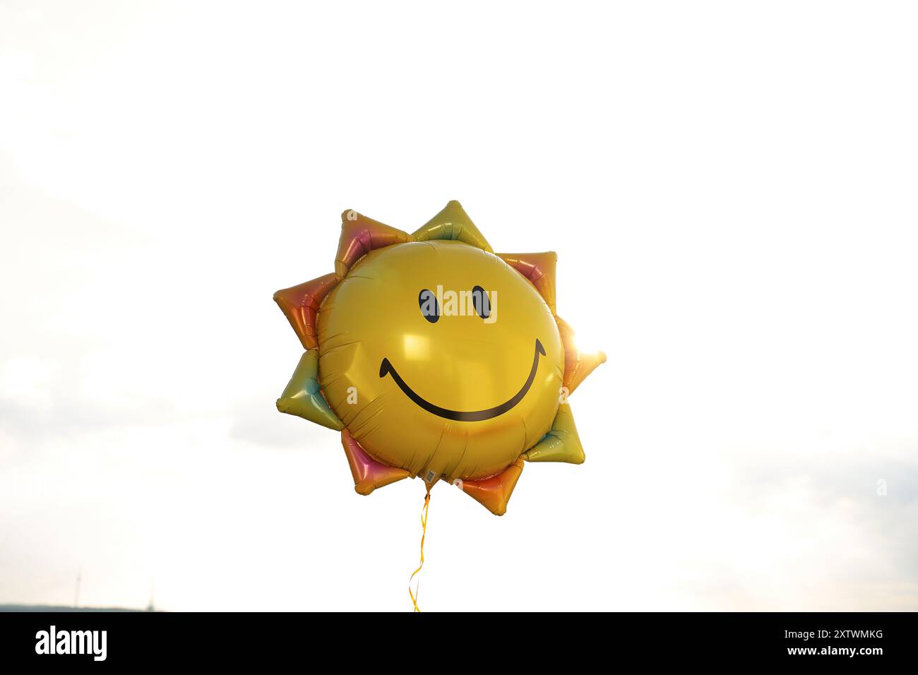 Un pallone all'elio sorridente a forma di sole con un vibrante colore giallo galleggia sullo sfondo di un cielo luminoso. Foto Stock