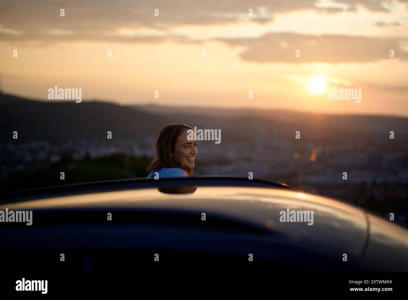 Donna sorridente che gode di una vista panoramica del tramonto dal tetto di un'auto con colline sullo sfondo. Foto Stock