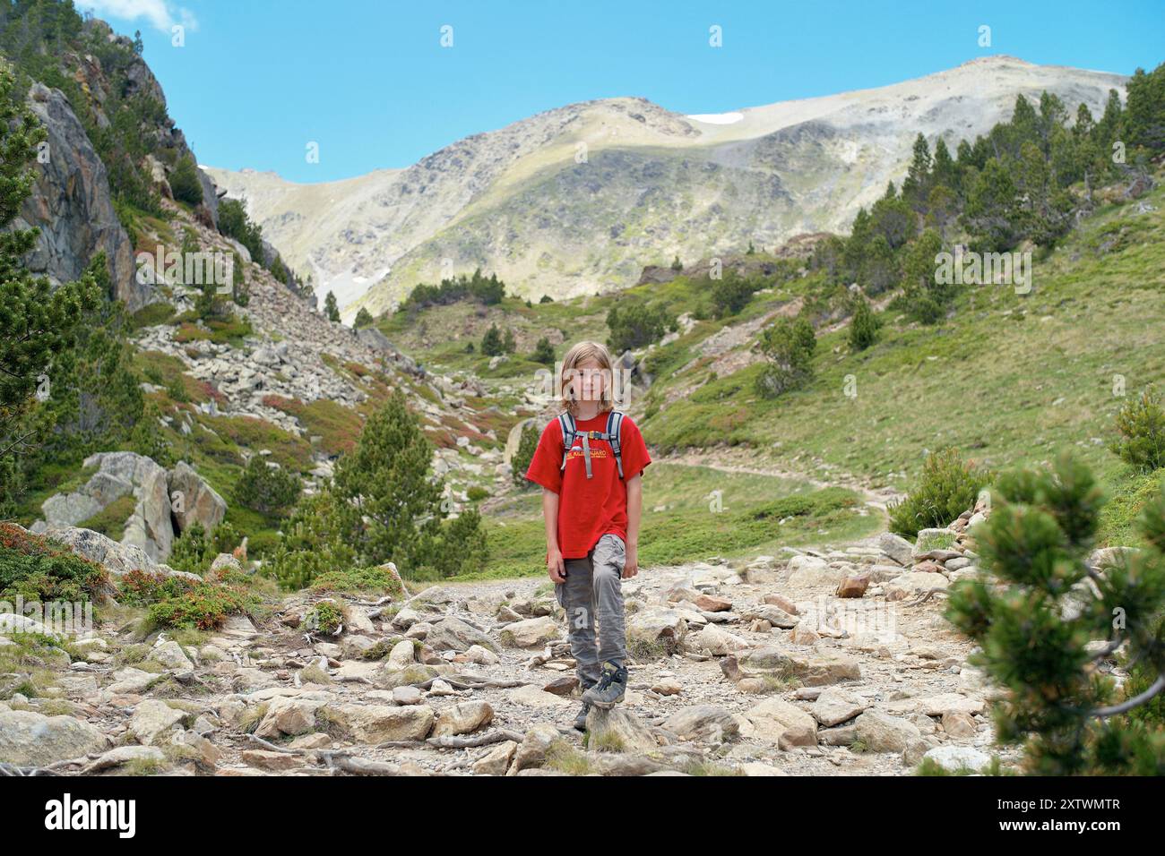 Giovane escursionista con uno zaino rosso che cammina su un sentiero roccioso di montagna circondato da una vegetazione lussureggiante. Foto Stock