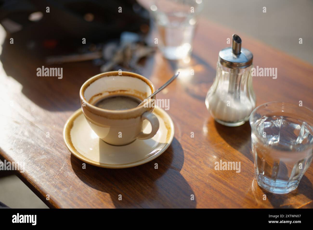 Caffè mattutino in una tazza bianca con piattino, accompagnato da un distributore di zucchero e un bicchiere d'acqua, su un tavolo di legno inondato dalla luce del sole. Foto Stock