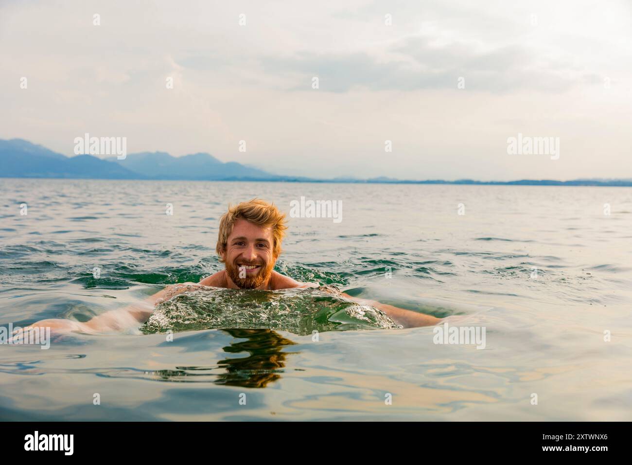 Un uomo sorridente barbuto che nuota in un lago azzurro con montagne sullo sfondo sotto un cielo nuvoloso. Foto Stock