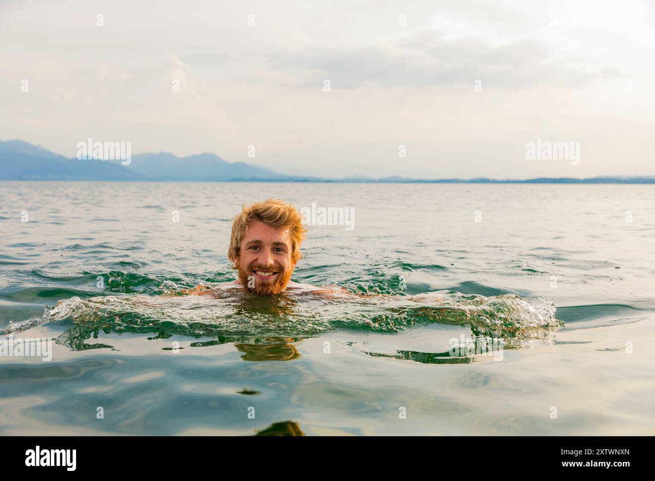 Uomo sorridente che nuota in un lago tranquillo con montagne in lontananza al tramonto. Foto Stock