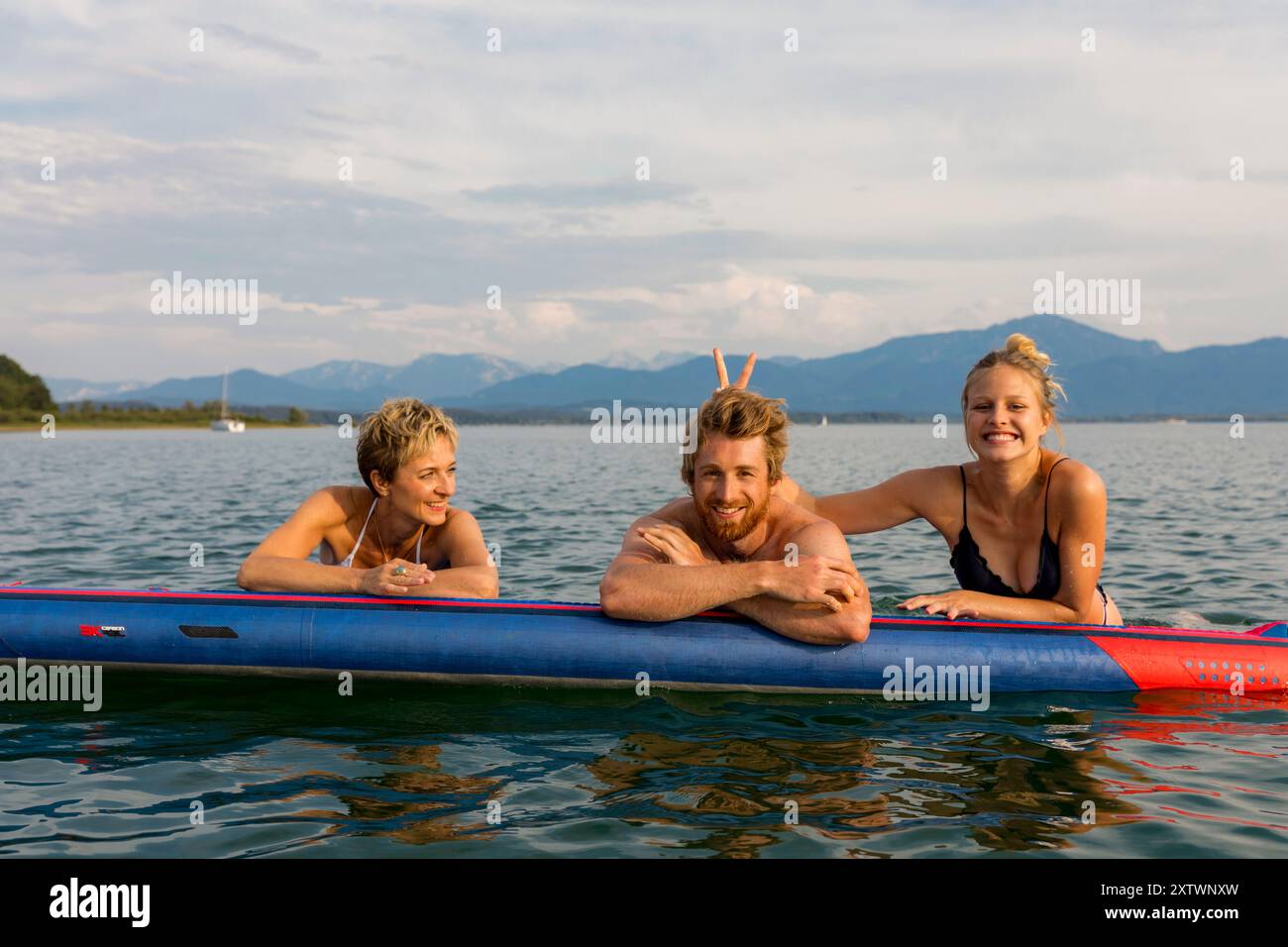 Tre amici sorridenti su un kayak blu in un lago con montagne sullo sfondo. Foto Stock