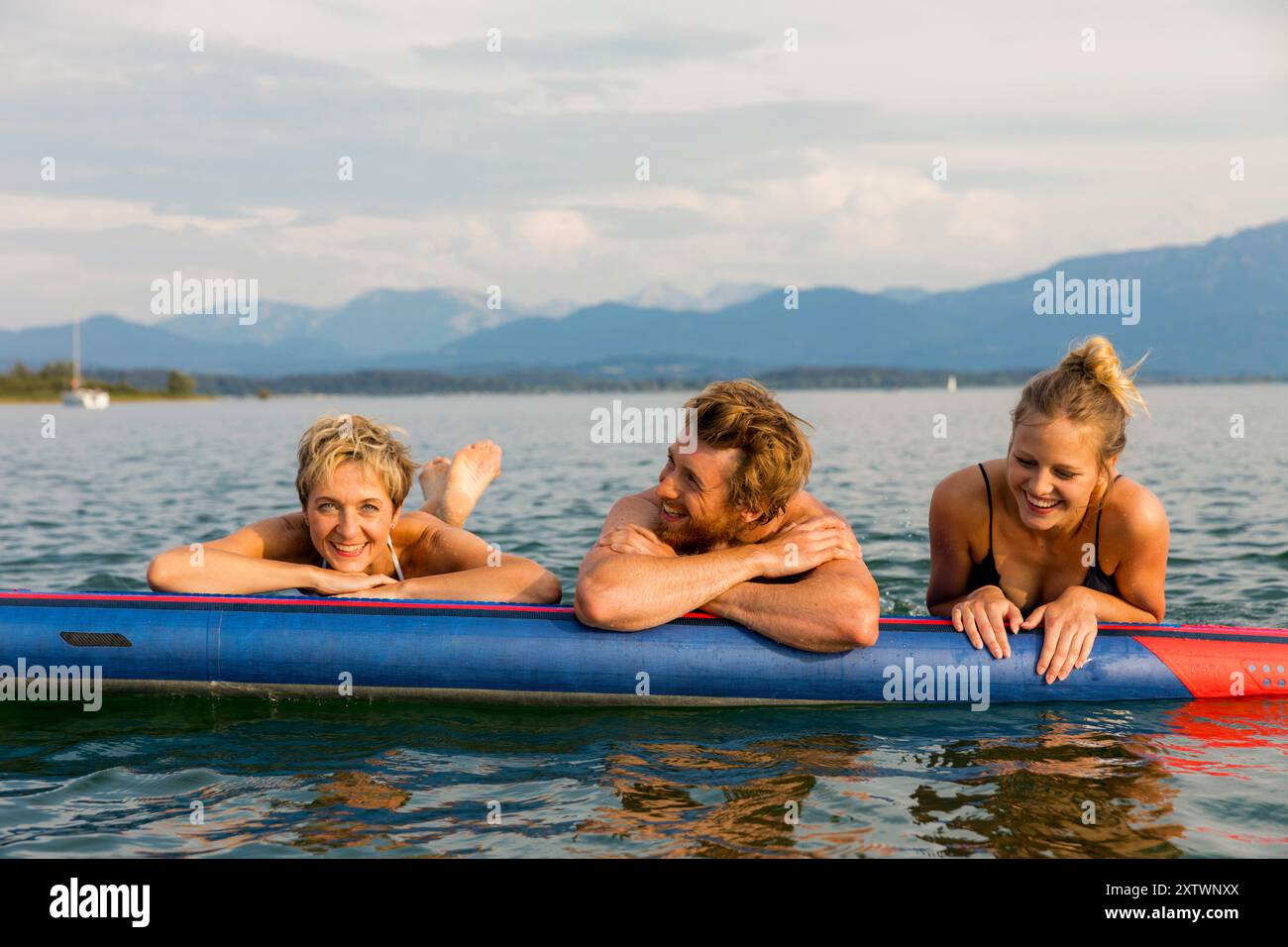 Tre amici sorridono e si rilassano su un paddleboard in un lago con montagne sullo sfondo in una giornata di sole. Foto Stock