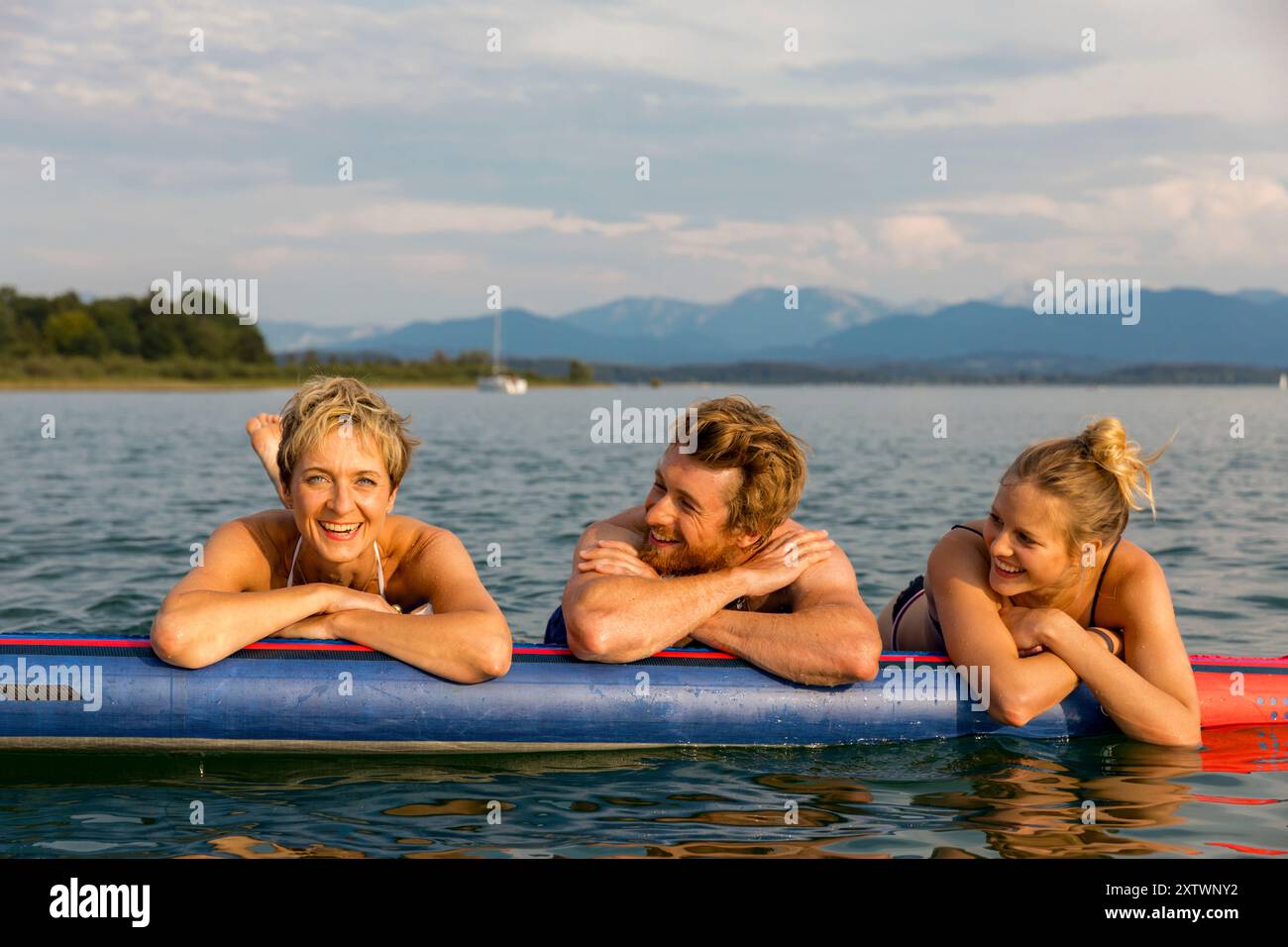 Tre amici si godono una giornata di sole su un paddleboard in un lago calmo con montagne in lontananza. Foto Stock
